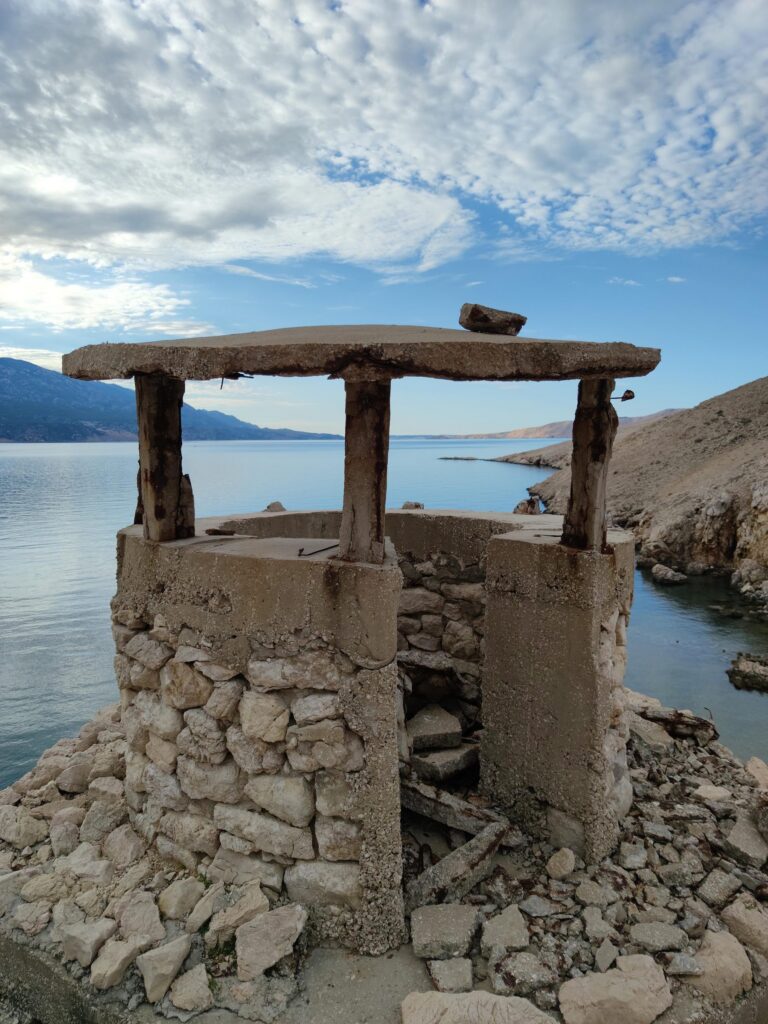 Kleines Steinhaus mit Holzdach über einer Zisterne, im Hintergrund das Meer