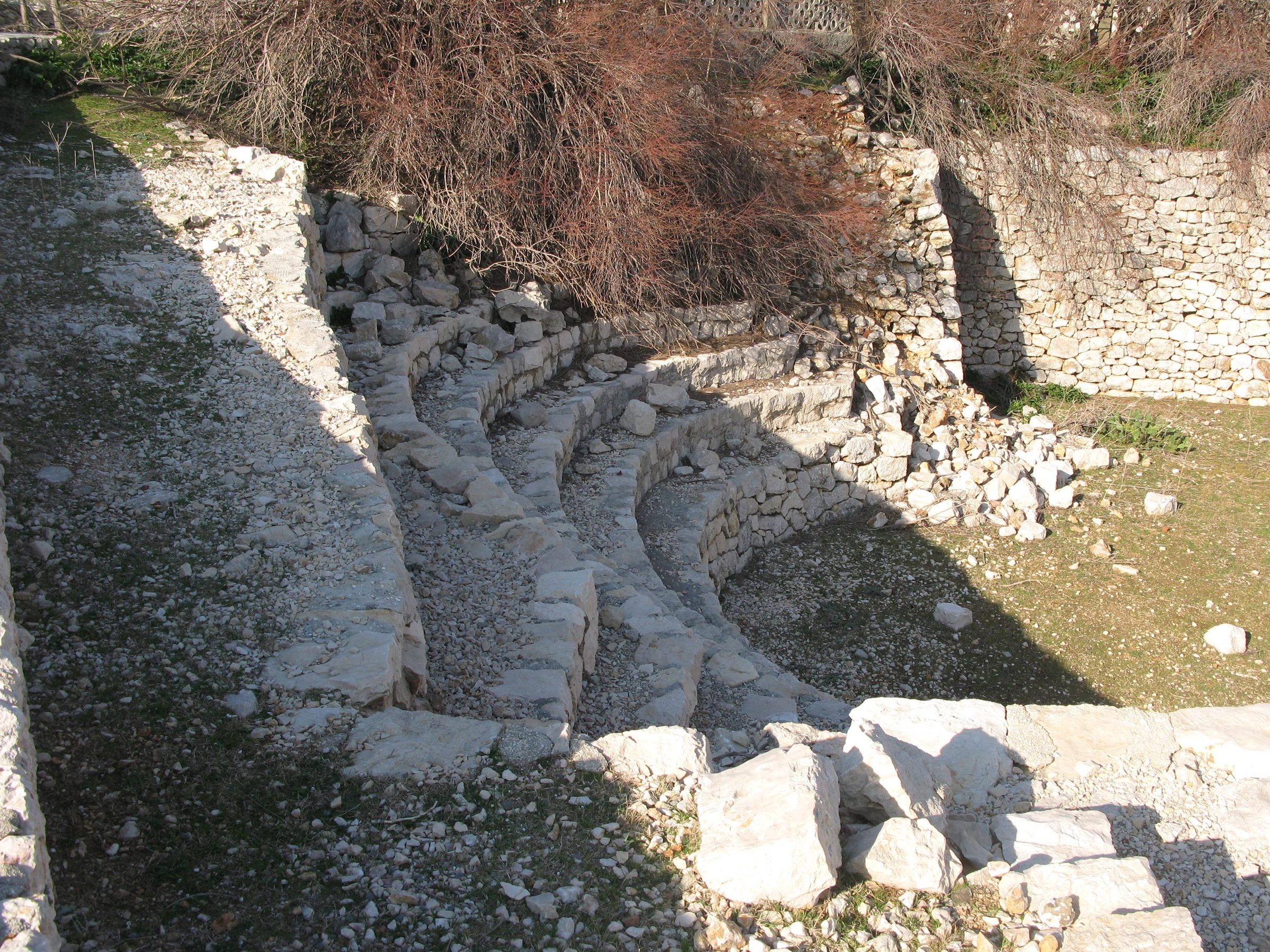 Runde, mit Trockenmauern eingefasste, grasbewachsene Grube